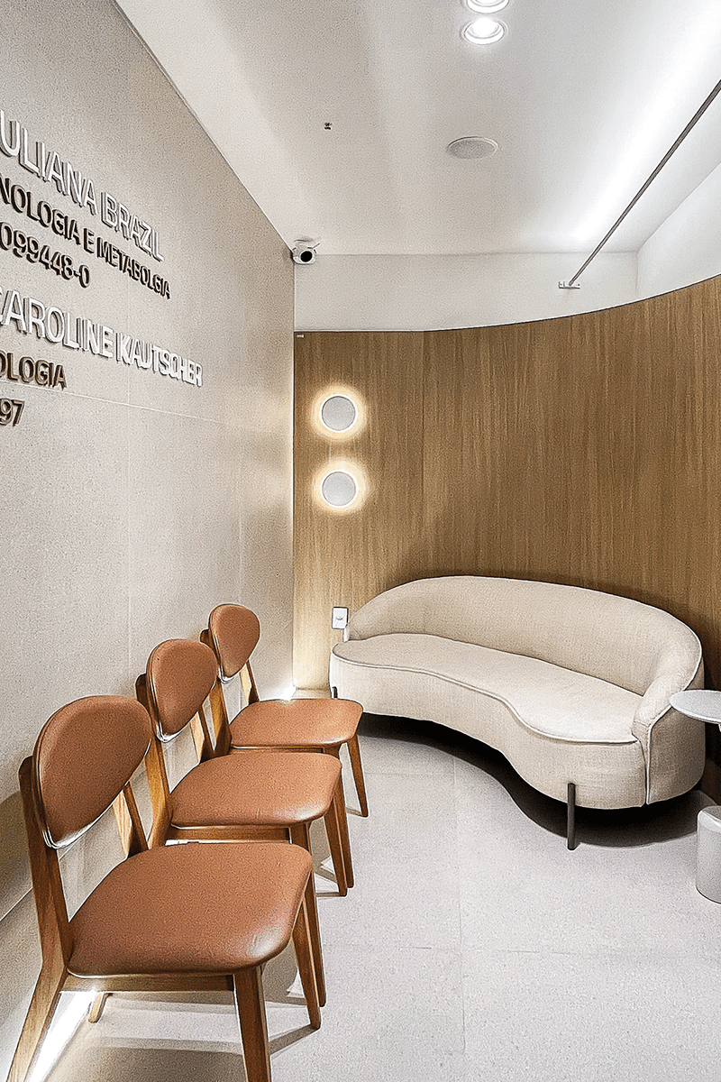 Na recepção, o painel amadeirado curvo se adequou ao sofá e não foi levado até o teto para trazer mais leveza. Alguns detalhes de iluminação foram destaque, como a luminária skyline, o perfil de led no piso atrás das cadeiras e a dupla de arandelas com iluminação indireta